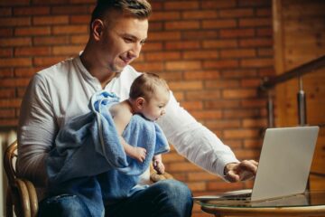Vader is op computer bezig met verlof regelen terwijl hij baby in zijn armen houdt