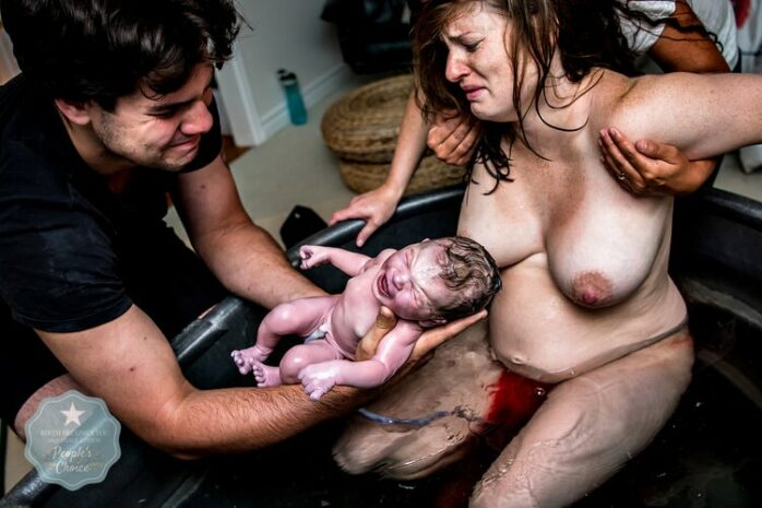 Man houdt pasgeboren baby huilend vast, met vrouw huilend op de achtergrond