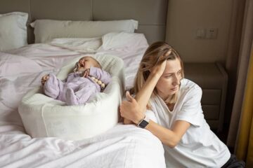 Moeder met postnatale depressie hangt tegen het bed aan terwijl baby op bed ligt te huilen