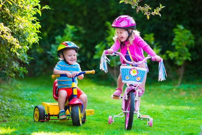 Peuter speelt op driewieler met bak, samen met oudere zus op kinderfiets met zijwieltjes