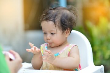 Meisje zit buiten in kinderstoel en ontdekt voedsel met haar handen, passend bij het voedingsschema van een baby van 8 maanden