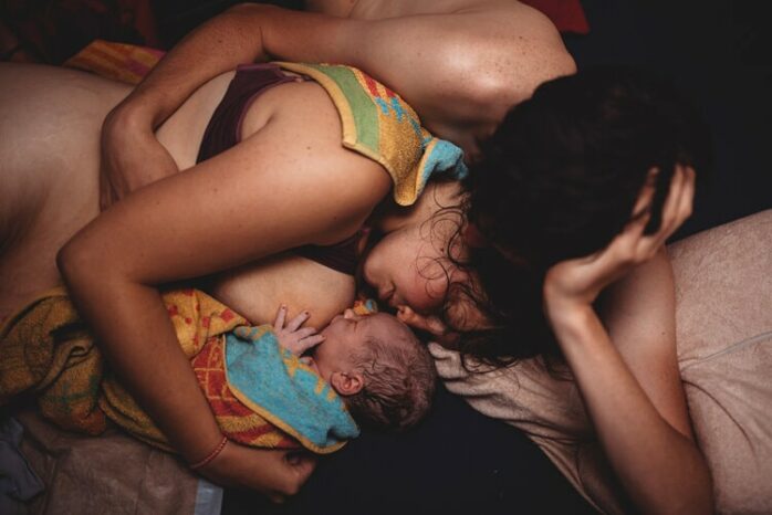 Vrouw ligt uitgeput op haar zij met haar baby aan de borst, terwijl haar partner haar knuffelt