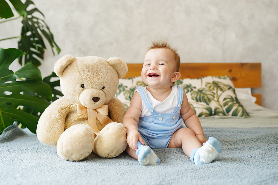 25 Wochen altes Baby sitzt neben einem Teddy und lacht