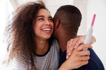 Frau fällt ihrem Partner strahlend um den Hals mit einem positiven Schwangersachftstest in der Hand