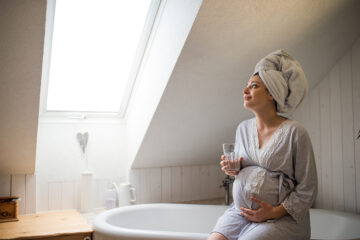 Verstopfung in der Schwangerschaft - Schwangere sitzt auf dem Rand der Wanne und trinkt ein Glas Wasser
