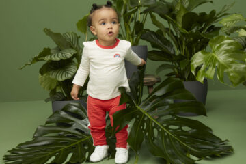 Baby steht neben einer Pflanze und trägt Babykleidung aus Bio-Baumwolle.