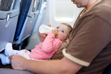 Reise mit Baby: Papa hat sein Baby auf dem Schoß im Flugzeug und füttert es mit einer Flasche.